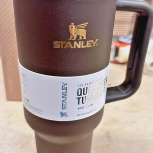May include: A brown Stanley Quencher tumbler with a gold-coloured rim and handle. The tumbler has the Stanley logo and the text "Quencher Tumbler" printed on it. It holds 1.18 L.