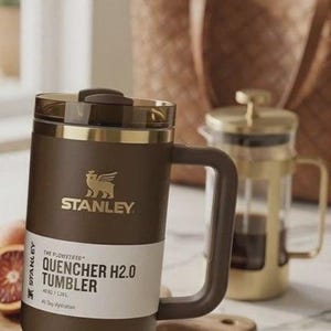 May include: A brown Stanley Quencher H2.0 tumbler with a handle, gold accents, and the Stanley logo. The tumbler is on a marble surface, with a French press and a wooden cutting board in the background. The tumbler is 1.18 L.
