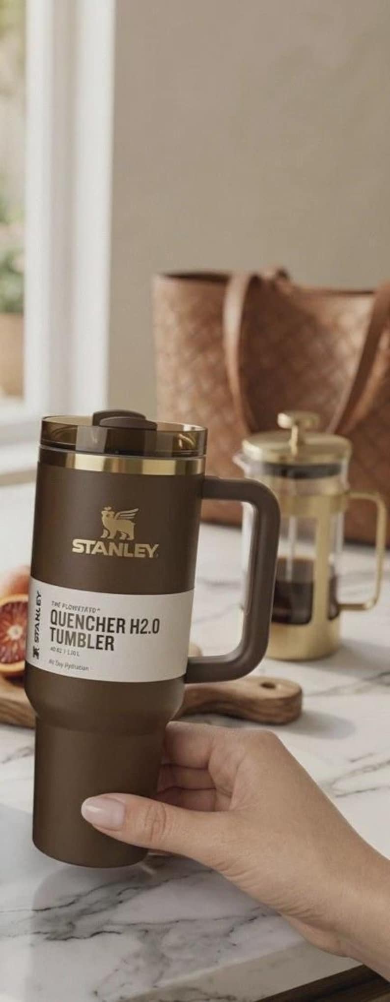 May include: A brown Stanley Quencher H2.0 tumbler with a handle, gold accents, and the Stanley logo. The tumbler is on a marble surface, with a French press and a wooden cutting board in the background. The tumbler is 1.18 L.