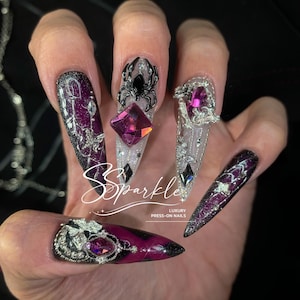 May include: Long stiletto press-on nails featuring a deep purple and black glitter base with intricate silver filigree, 3D spider charms, and faceted magenta crystals. The text Sparkle Luxury Press-On Nails is printed in white across the center.