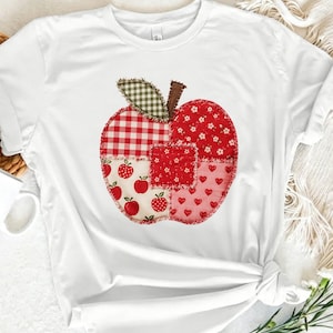 May include: A white short-sleeve t-shirt featuring a large apple-shaped graphic on the front. The apple is composed of various fabric-style patches in red, white, and green, including patterns of gingham, small flowers, hearts, and tiny apples.