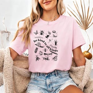 Może przedstawiać: Jasnoróżowy t-shirt z krótkim rękawem z czarnym nadrukiem graficznym przedstawiającym różne owady oraz napis "be kind to all kinds" ułożony w zakrzywionej linii. Koszulka ma okrągły dekolt i jest zestawiona z jasnoniebieskimi szortami dżinsowymi.