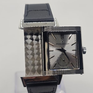 Puede incluir: Un reloj rectangular de tono plateado con correa de cuero negro. La esfera plateada con efecto rayos de sol presenta marcadores de hora plateados, manecillas negras, una subesfera y el texto Jaeger-LeCoultre. La caja tiene un panel lateral metálico.
