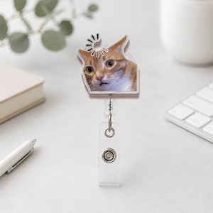 May include: A retractable badge reel featuring a surprised orange tabby cat face. The cat's image is set against a white background, with a black and white swirl design above its head. The reel has a clear plastic clip and a metal snap.