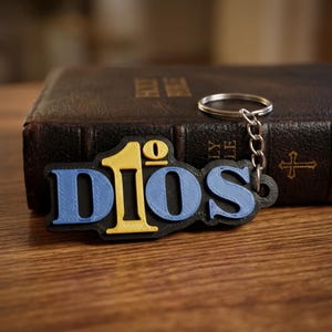 May include: A keychain featuring the word DIOS in a layered design. The letters D and S are light blue, the number 1 is yellow, and the letter I is yellow with a small zero above it. The entire word is set against a black base with a metal ring.