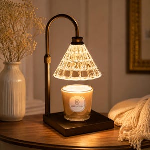 Op de afbeelding: Een kaarsenverwarmingslamp met een heldere, getextureerde glazen kap en een zwart metalen frame. De lamp staat op een vierkante basis, met een brandende kaars in een glazen pot eronder. De kaarsenpot heeft een label met "MANYTNOW".