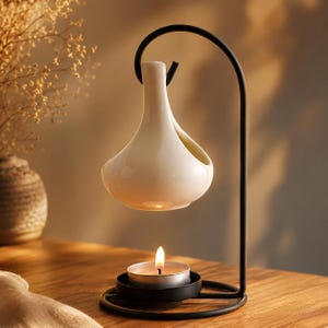 May include: A white ceramic oil burner with a teardrop-shaped vessel suspended from a black metal stand. A small tea light candle sits in a circular black metal base directly beneath the vessel, providing heat to the ceramic container above.