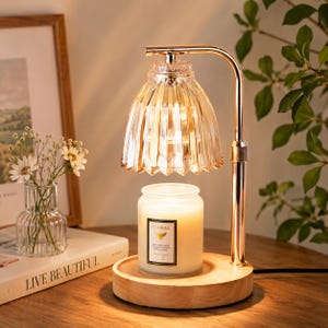 May include: A candle warmer with a gold-coloured frame, glass shade, and wooden base. A white candle with a floral label sits on the base. A small vase of flowers and a framed picture are in the background.