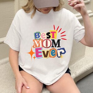 Puede incluir: Camiseta blanca con el texto "Best Mom Ever" en letras coloridas de estilo recortado. El diseño incluye una estrella y rayos rojos. Una prenda casual para el uso diario.