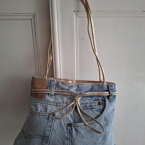 Op de afbeelding: Een lichtblauwe denim tote bag met goudkleurige bandjes en accenten. De tas is gemaakt van hergebruikte jeans, met zakken en een gouden strikdetail. De tas hangt tegen een witte deur.