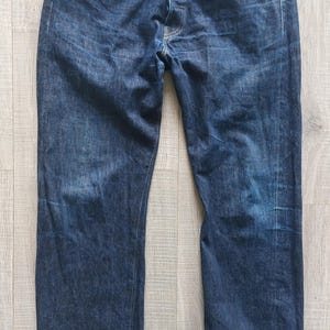 May include: Dark blue denim jeans with a button fly, five-pocket design, and a straight leg. The jeans have a classic, worn look with subtle fading and creasing. The cuffs are rolled up, revealing the selvedge.