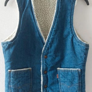 Può includere: Un gilet di jeans blu con fodera in sherpa color crema e due tasche frontali. Il gilet ha una chiusura con bottoni e una piccola etichetta rossa Levi's su una tasca. Il gilet è appeso a una gruccia nera.