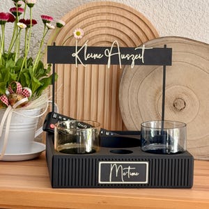 Könnte beinhalten: Ein schwarzer Holz-Organizer mit zwei Glashaltern, einem Fernbedienungsfach und dem Text "Me time". Der Organizer hat ein Schild mit der Aufschrift "Kleine Auszeit". Ein kleiner Blumentopf und Holzdekor befinden sich im Hintergrund.