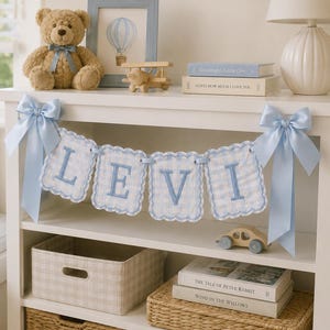 May include: A decorative banner featuring four scalloped, light blue and white gingham fabric panels, each embroidered with a letter to spell LEVI. The banner is accented with large, light blue satin bows on both ends, hanging from a white shelf.