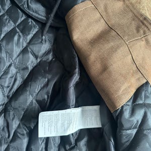 May include: Close-up of a brown jacket with a black quilted lining. The jacket has a hood and a label with text. The outer material is a textured brown fabric.