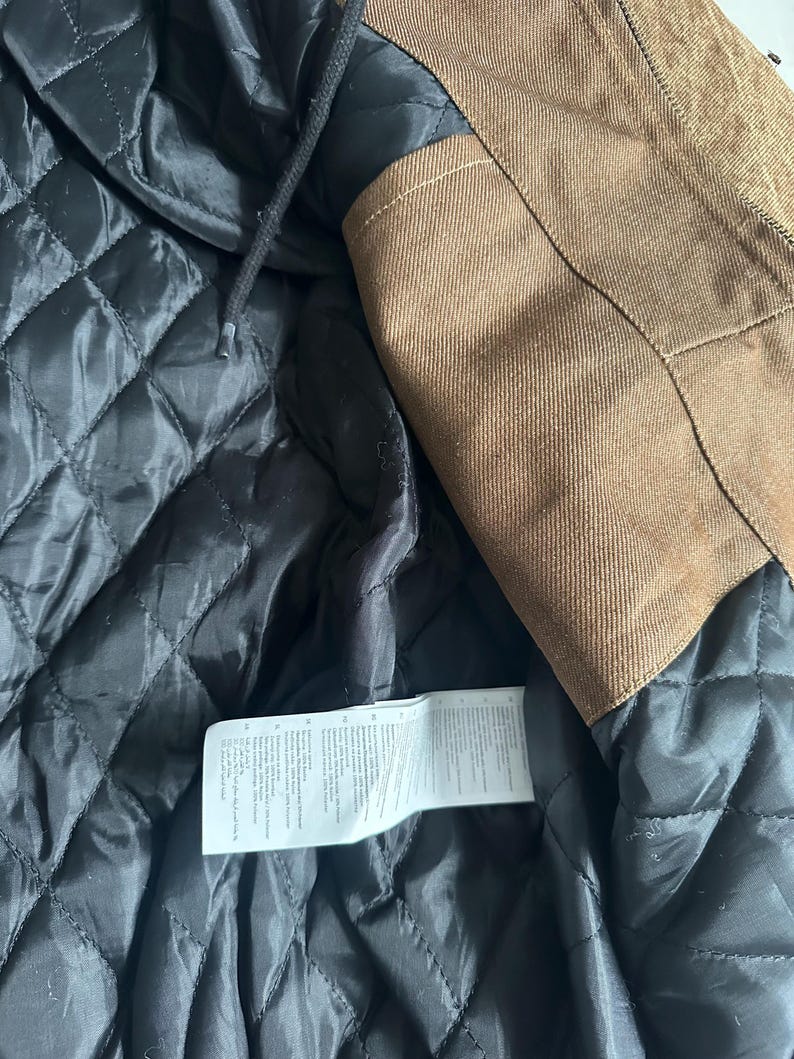May include: Close-up of a brown jacket with a black quilted lining. The jacket has a hood and a label with text. The outer material is a textured brown fabric.