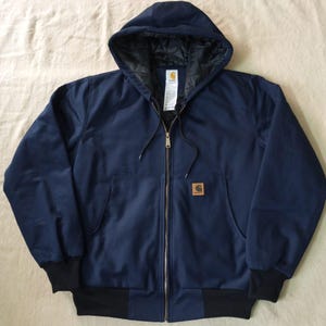 Puede incluir: Una chaqueta con capucha y cremallera en color azul marino, con forro acolchado negro, cierre frontal de cremallera y puños y dobladillo acanalados en negro. Un pequeño parche cuadrado de color tostado con el logo de Carhartt en el bolsillo.