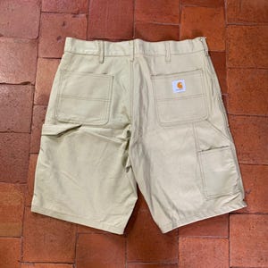 Könnte beinhalten: Beige Arbeitsshorts mit strukturiertem Bund und Gürtelschlaufen, zwei großen Gesäßtaschen und einer Werkzeugtasche am rechten Bein. Ein kleines rechteckiges Carhartt-Markenetikett mit orangefarbenem und weißem Text ist auf die rechte Gesäßtasche genäht.