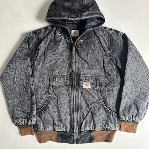 May include: A black and gray hooded jacket with a zip-up front and brown ribbed cuffs and hem. The jacket features a Carhartt logo patch on the front pocket. The jacket has a distressed, acid-wash appearance.