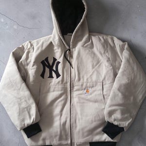 May include: A beige hooded jacket with a black lining and a black zipper. The jacket features the New York Yankees logo on the chest and a Carhartt logo patch. The cuffs and hem are black.