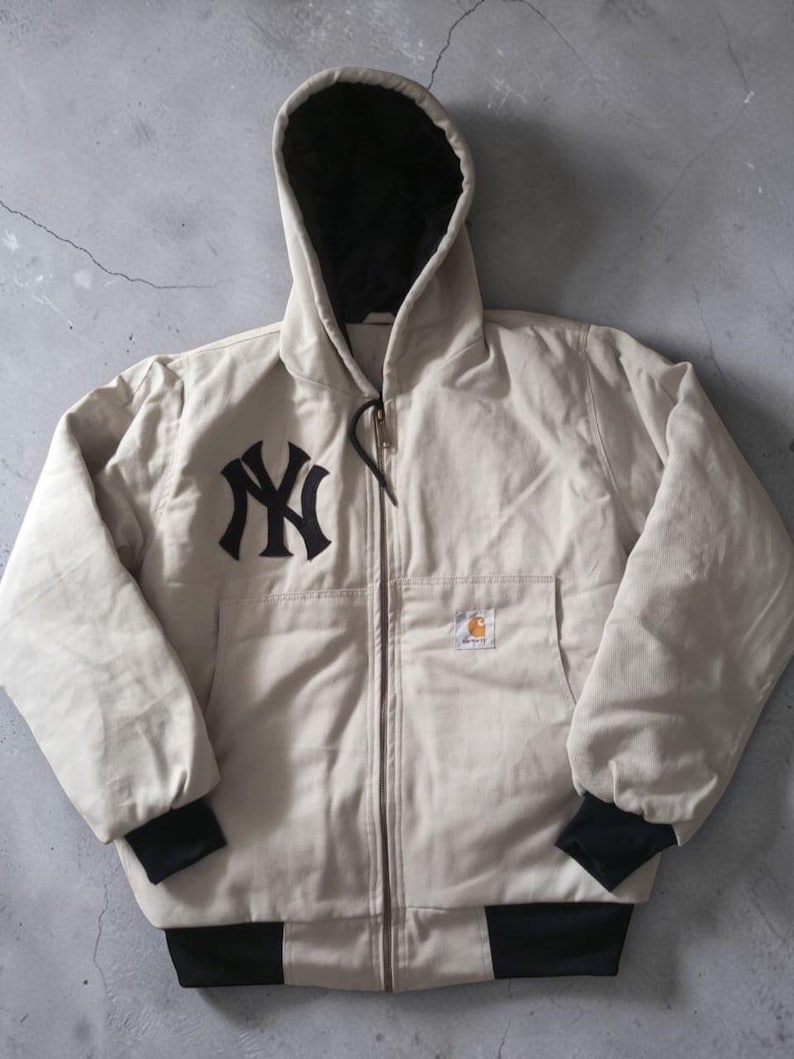 May include: A beige hooded jacket with a black lining and a black zipper. The jacket features the New York Yankees logo on the chest and a Carhartt logo patch. The cuffs and hem are black.
