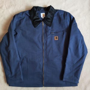 May include: A navy blue jacket featuring a black collar, a full-length front zip, and long sleeves. A small rectangular brown patch with the Carhartt logo is positioned on the left chest, just below a horizontal zipped pocket. A white brand label is visible at the inner neck.