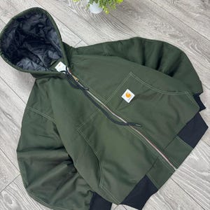 Pode incluir: Um casaco com capuz em verde-azeitona escuro com fecho frontal completo e punhos e bainha canelados em preto. O casaco apresenta um forro interior acolchoado preto, um capuz com cordão e um emblema quadrado da marca Carhartt no bolso.