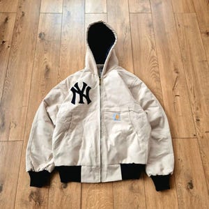 Op de afbeelding: Beige jas met capuchon en het New York Yankees-logo in zwart en marineblauw op de borst. De jas heeft een zwarte geribbelde zoom en manchetten, een ritssluiting en een marineblauwe voering. Gemaakt van duurzaam materiaal.