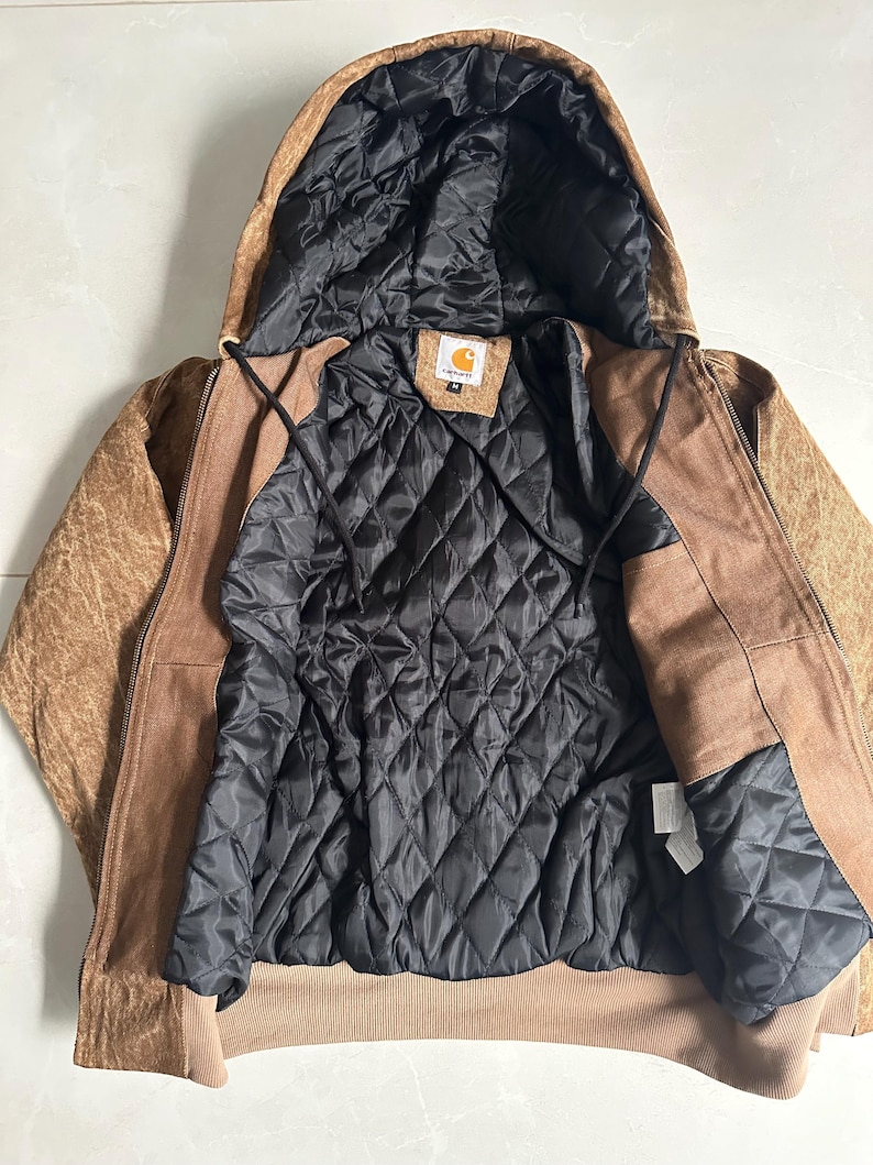 May include: A brown hooded jacket with a black quilted lining. The jacket has a zipper closure and a brown exterior with a textured appearance. The interior lining is black and quilted. The jacket has a tag with a logo.