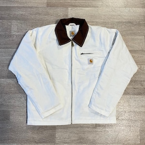 May include: A white zip-up jacket featuring a dark brown fold-down collar, long sleeves, and a silver-toned metal zip closure. A small rectangular Carhartt brand patch is visible on the left chest pocket area, which includes a horizontal zip.