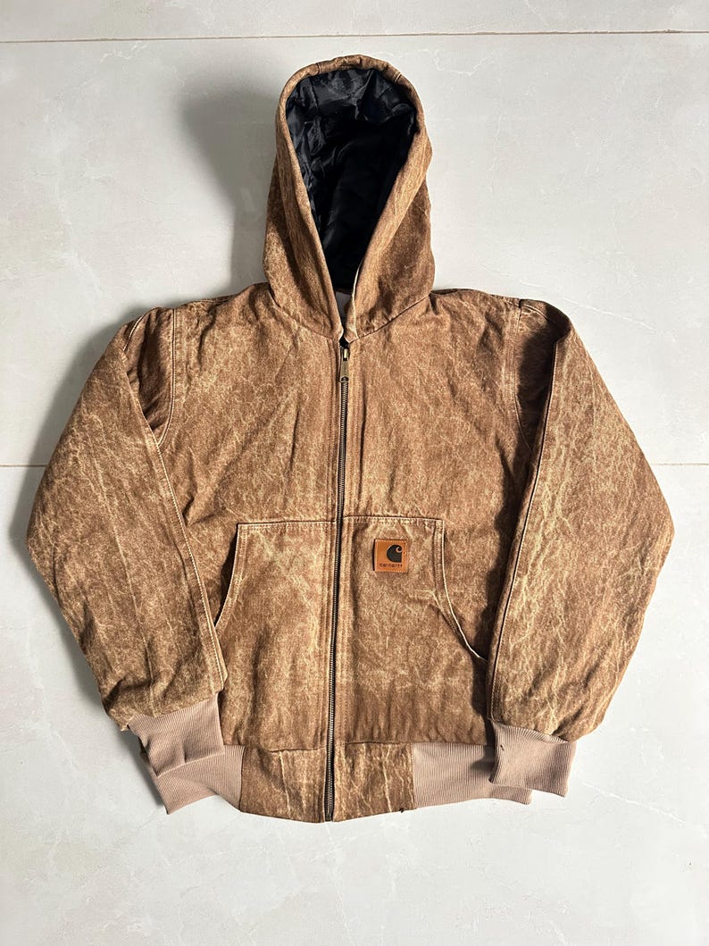 May include: Brown hooded jacket with a zip-up front, a front pocket, and ribbed cuffs and hem. The jacket features a small square logo patch on the front. The interior lining of the hood is black.