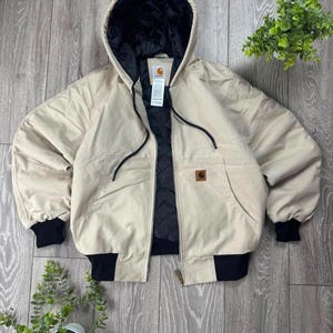 Puede incluir: Una chaqueta beige con capucha, forro interior acolchado negro, puños acanalados negros y dobladillo acanalado negro. La chaqueta cuenta con cierre de cremallera frontal, cordones negros para la capucha y un parche cuadrado de la marca Carhartt.