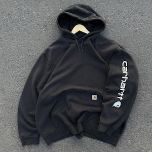 May include: Black hooded sweatshirt with a kangaroo pocket and the Carhartt logo on the sleeve. The sweatshirt is hanging on a wooden hanger. The Carhartt logo is also on the front pocket.