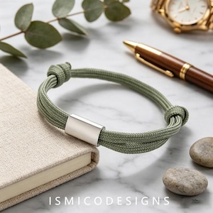 May include: A sage green cord bracelet featuring a smooth, rectangular silver-toned metal bead at the centre. The bracelet is constructed from two parallel strands of woven cord, secured with adjustable sliding knots on each side of the metal bead.