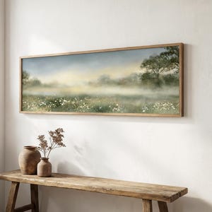 Puede incluir: Una pintura de paisaje panorámico en un marco de madera que muestra un prado brumoso con flores silvestres blancas, follaje verde suave y árboles distantes bajo un cielo nublado y tenue. La obra de arte se exhibe sobre un banco de madera rústico.