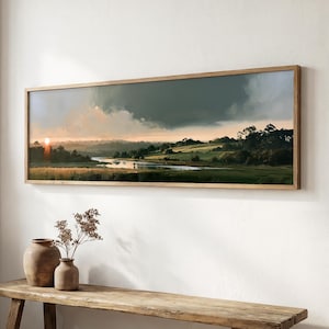 Op de afbeelding: Een panoramisch landschapsschilderij in een dunne houten lijst met een rivier die door groene velden slingert onder een dramatische, donkere, bewolkte lucht bij zonsondergang, waarbij de zon zichtbaar is aan de horizon op een witte muur.