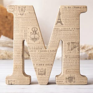 Op de afbeelding: Een grote houten letter "M" met een vintage krantenprint ontwerp. De letter is beige met zwarte tekst en illustraties, waaronder de Eiffeltoren. Ideaal voor woondecoratie of gepersonaliseerde cadeaus.