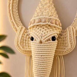 May include: A cream-colored macrame wall hanging shaped like an elephant head. The design features intricate knotted cord patterns, a circular frame, smooth round beads, and clear faceted crystal-like accents hanging from the ears and trunk.