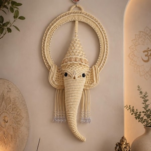 May include: A cream-colored macrame wall hanging shaped like the head of Ganesha, featuring a circular frame, intricate knotted cord details, a pointed crown adorned with clear beads, and two black eyes, suspended by a small circular hook.