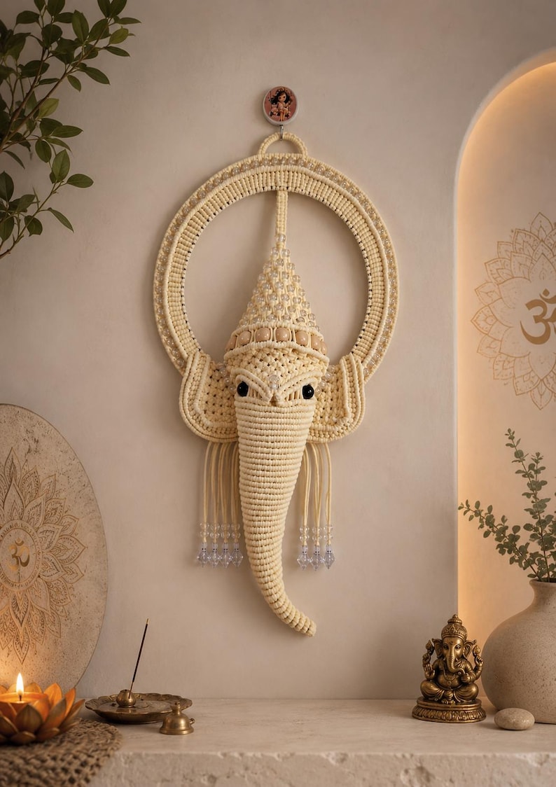 May include: A cream-colored macrame wall hanging shaped like the head of Ganesha, featuring a circular frame, intricate knotted cord details, a pointed crown adorned with clear beads, and two black eyes, suspended by a small circular hook.