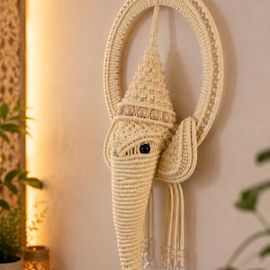 May include: A cream-colored macrame wall hanging shaped like an elephant head, featuring a circular frame, a pointed crown, wooden beads, and clear crystal-like accents. The intricate knotted cord design forms the trunk, ears, and head.