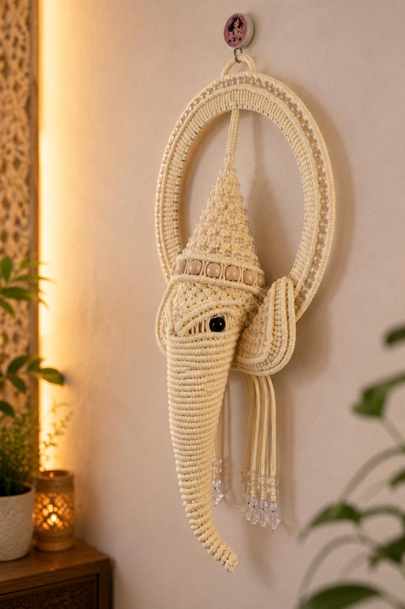 May include: A cream-colored macrame wall hanging shaped like an elephant head, featuring a circular frame, a pointed crown, wooden beads, and clear crystal-like accents. The intricate knotted cord design forms the trunk, ears, and head.