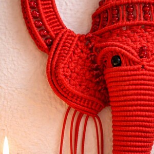 May include: A bright red macrame wall hanging shaped like an elephant head, featuring intricate knotted cord patterns, embedded red beads, a prominent trunk, large ears, and three dangling red cord accents with small red bead drops at the ends.