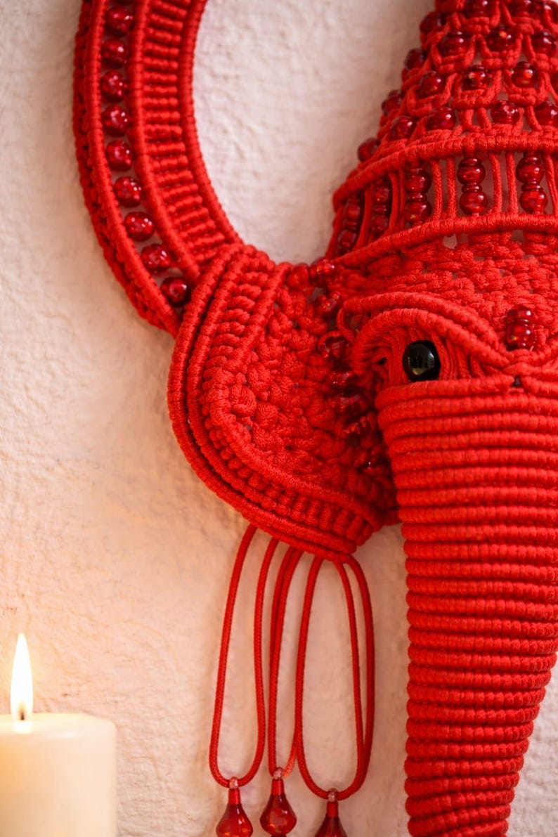 May include: A bright red macrame wall hanging shaped like an elephant head, featuring intricate knotted cord patterns, embedded red beads, a prominent trunk, large ears, and three dangling red cord accents with small red bead drops at the ends.