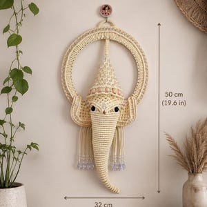 May include: A cream-colored macrame wall hanging shaped like an elephant head with a circular frame, adorned with clear beads and wooden accents. The piece measures 19.6 inches in height and 12.6 inches in width, featuring intricate knotted cord details.