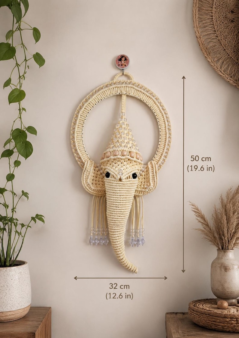 May include: A cream-colored macrame wall hanging shaped like an elephant head with a circular frame, adorned with clear beads and wooden accents. The piece measures 19.6 inches in height and 12.6 inches in width, featuring intricate knotted cord details.