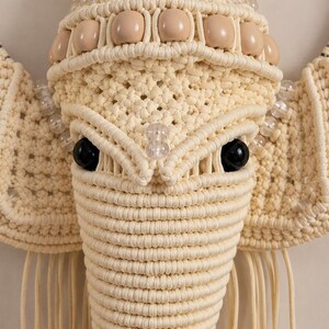 May include: A macrame wall hanging shaped like an elephant head, crafted from cream-colored cord. The design features large ears, a trunk, and prominent black eyes. It is decorated with rows of clear glass beads and smooth beige wooden beads.