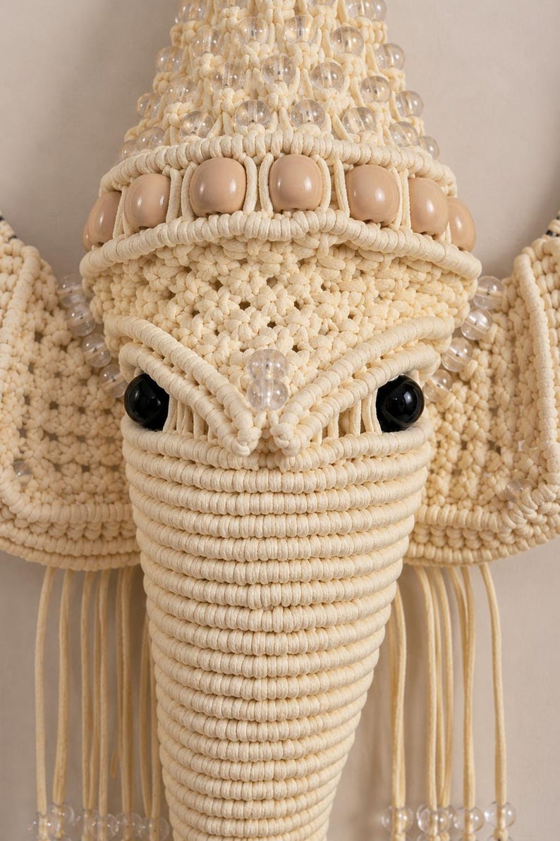 May include: A macrame wall hanging shaped like an elephant head, crafted from cream-colored cord. The design features large ears, a trunk, and prominent black eyes. It is decorated with rows of clear glass beads and smooth beige wooden beads.