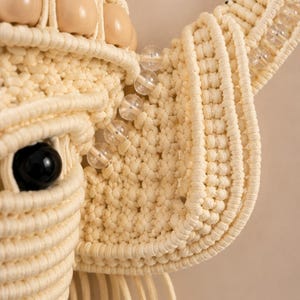 May include: A macrame wall hanging shaped like an animal head, crafted from cream-colored cord. The design features a prominent black bead for an eye, smooth beige wooden beads, and clear glass beads accenting the ear and forehead structure.