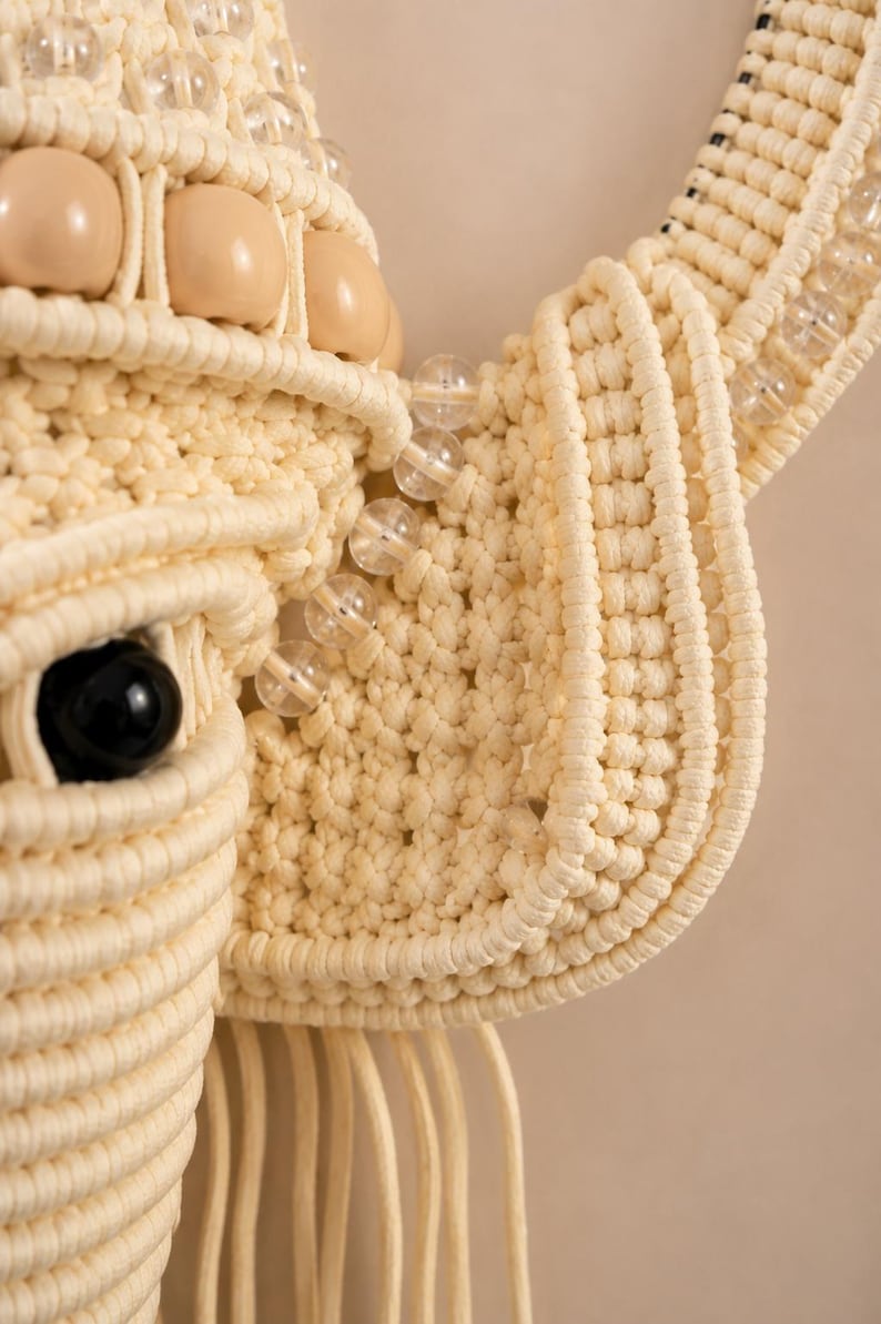 May include: A macrame wall hanging shaped like an animal head, crafted from cream-colored cord. The design features a prominent black bead for an eye, smooth beige wooden beads, and clear glass beads accenting the ear and forehead structure.
